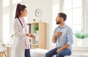 Young man sitting on the couch in the doctor’s office and pointing to his chest to the doctor during medical examination in clinic. Physician listening to the patient’s complaints.