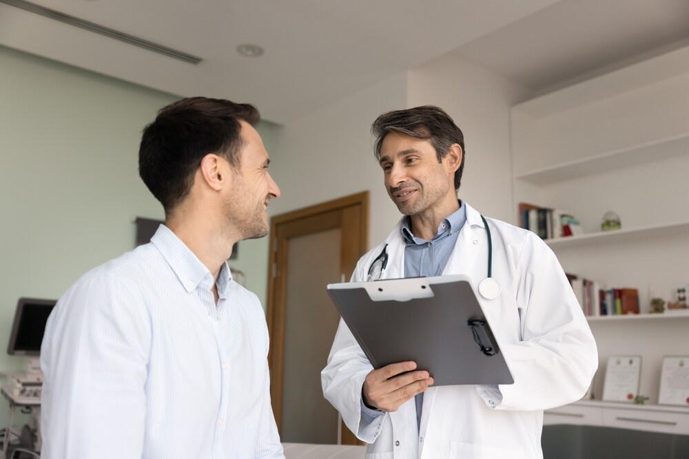 Positive optimistic Latin doctor giving consultation, recommendation to patient after checkup, talking to man in examination room, holding pad with medical records, writing notes, filling out survey