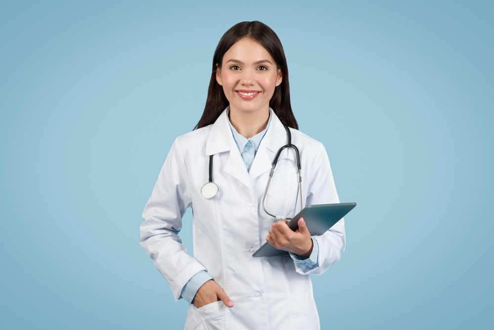 Confident young female doctor with stethoscope around her neck holding digital tablet, ready for patient care, against blue background