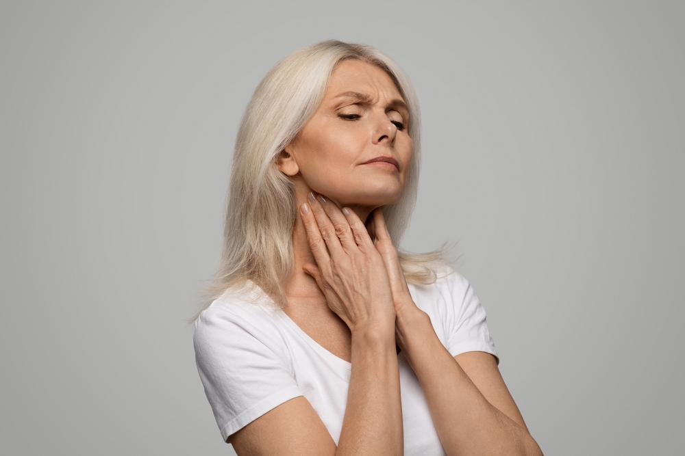 Portrait of unhappy sad sick mature woman suffering from sore throat, ill senior lady touching neck with hands, feeling ache, standing isolated over grey studio background, copy space